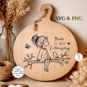 Puede incluir: Una tabla de cortar redonda de madera con un asa y una ilustraci&oacute;n decorativa. La ilustraci&oacute;n presenta a una ni&ntilde;a sentada en una rama con un p&aacute;jaro y una mariposa. El texto en la tabla dice "Heute ist mein Lieblingstag".