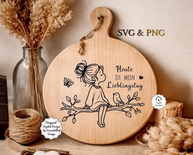 Puede incluir: Una tabla de cortar redonda de madera con un asa y una ilustraci&oacute;n decorativa. La ilustraci&oacute;n presenta a una ni&ntilde;a sentada en una rama con un p&aacute;jaro y una mariposa. El texto en la tabla dice "Heute ist mein Lieblingstag".