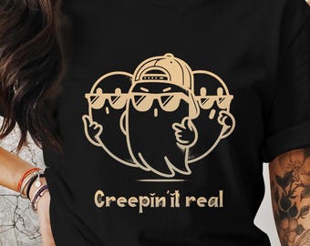 May include: Black t-shirt with a tan graphic of three cartoon ghosts wearing sunglasses and a baseball cap. The phrase "Creepin' it real" is printed below the ghosts. The bottom of the image includes the text "PNG & SVG WITHOUT text incl."