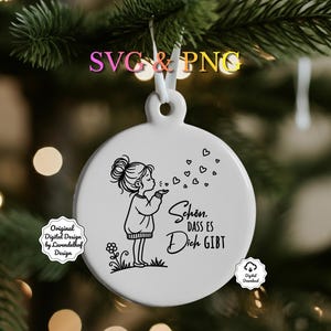 Puede incluir: Un adorno navide&ntilde;o de cer&aacute;mica blanca con una ilustraci&oacute;n en blanco y negro de una ni&ntilde;a soplando burbujas en forma de coraz&oacute;n. El adorno tiene el texto "Sch&ouml;n, dass es Dich gibt". Cuelga de una cinta blanca.