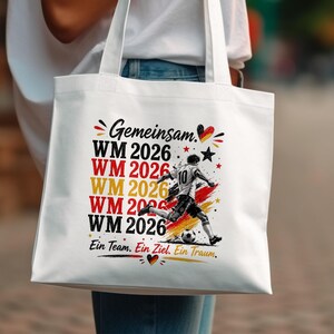 May include: White tote bag featuring a soccer player kicking a ball, with the text "Gemeinsam WM 2026" in red, yellow, and black. The bag also includes the phrase "Ein Team. Ein Ziel. Ein Traum."