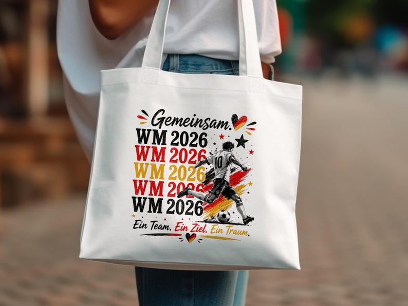 May include: White tote bag featuring a soccer player kicking a ball, with the text "Gemeinsam WM 2026" in red, yellow, and black. The bag also includes the phrase "Ein Team. Ein Ziel. Ein Traum."