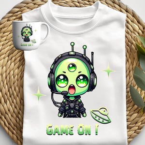 Könnte beinhalten: Weißes Sweatshirt und Becher mit einem Cartoon-Alien mit drei Augen, das ein Headset und eine Uniform trägt. Das Alien ist grün mit großen Augen und der Text "GAME ON!" wird auf beiden Artikeln angezeigt. Eine grüne Untertasse ist ebenfalls abgebildet.