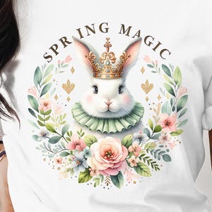 Könnte beinhalten: Weißes T-Shirt mit einer Aquarell-Illustration eines Kaninchens, das eine goldene Krone trägt und von Blumen umgeben ist. Der Text "SPRING MAGIC" steht über dem Kaninchen.