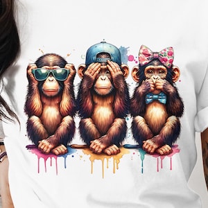 Puede incluir: Camiseta blanca con tres monos de dibujos animados en la pose 'No veo el mal, no oigo el mal, no hablo el mal'. Un mono lleva gafas de sol, otro una gorra azul y el tercero una pajarita floral. Cada mono tiene gotas de pintura de colores.