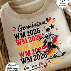 May include: Cream-colored sweatshirt featuring a soccer player kicking a ball, with the text "WM 2026" in red, yellow, and black. The design includes German text and a heart with the German flag colors. "Ein Team. Ein Ziel. Ein Traum."
