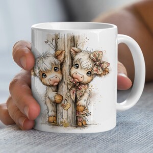 May include: White ceramic mug featuring a watercolor illustration of two adorable Highland cows peeking from behind a wooden post. The cows have fluffy white fur, brown ears, and are adorned with floral bows. A heart-shaped lock is on the post.