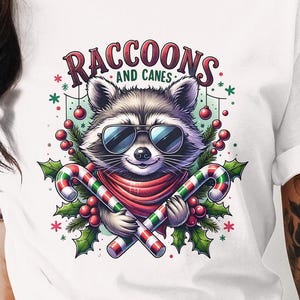 Könnte beinhalten: Ein weißes T-Shirt mit einem Cartoon-Waschbären, der eine Sonnenbrille und einen roten Schal trägt. Der Waschbär hält zwei Zuckerstangen. Der Text "Raccoons and Canes" befindet sich über dem Waschbären.