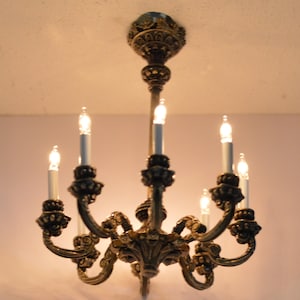 May include: An antique brass chandelier with six candle-style lights. The fixture has ornate, scrolling arms and decorative detailing. The bulbs are lit, casting a warm glow. A classic lighting fixture for a dining room or hallway.
