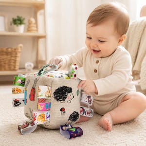 May include: A soft, beige fabric sensory cube toy featuring printed illustrations of hedgehogs and mushrooms. The cube is adorned with multiple colorful ribbon loops on its sides, each displaying various animal and nature-themed patterns.