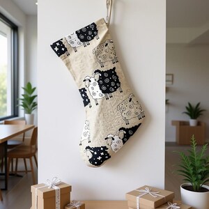 Könnte beinhalten: Ein beiger Weihnachtsstrumpf mit einem Muster aus schwarzen und weißen Schafen hängt an einer weißen Wand. Der Strumpf wird von einem goldfarbenen Haken gehalten. Mehrere braune Geschenkboxen mit weißem Band befinden sich auf einer Holzoberfläche.