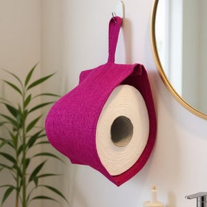 Könnte beinhalten: Ein magentafarbener Toilettenpapierhalter aus Filz mit einer Aufhängeschlaufe. Der Halter ist so geformt, dass er eine Rolle weißes Toilettenpapier aufnehmen kann. Das Badezimmerzubehör ist an einer weißen Wand montiert.