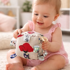 May include: A baby in a pink dress holds a soft, beige fabric cube toy with colorful floral and patterned accents. The toy has various fabric tags attached. The baby is sitting on a white carpet.