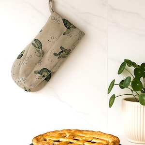 Puede incluir: Manopla de horno beige con un diseño de tortugas colgada en una pared blanca. El guante presenta tortugas verdes y burbujas. Una tarta horneada con una corteza de rejilla está en un plato oscuro debajo.