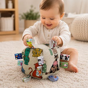 May include: A soft, beige fabric cube toy featuring a colourful print of various animals including cats and a cow. The cube has multiple small, patterned fabric loops attached to its edges, providing tactile sensory elements for play and exploration.