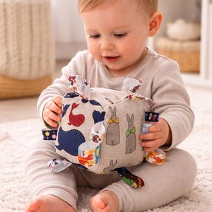 Puede incluir: Un cubo de juguete suave para bebés con diseños de ballenas y conejos. El cubo tiene cintas coloridas y lo sostiene un bebé con un body claro. El juguete está diseñado para el juego sensorial y el desarrollo temprano.