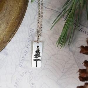 May include: A silver necklace with a rectangular pendant featuring a silhouette of a pine tree. The pendant is on a map with the words "Henness Branch" and "Barrel Springs Rd" visible.