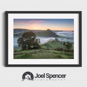 May include: A framed landscape photograph featuring a sunrise over rolling green hills partially covered in thick morning mist. A large tree stands in the foreground, while a prominent peak rises in the distance under a soft pink and blue sky.