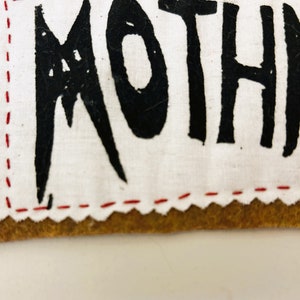 以下が含まれることがあります： 白い布のパッチのクローズアップ。黒いステッチと黒い塗料で書かれた「MOTHMAN」という言葉があります。パッチは、縁に赤いステッチが施された茶色の革の裏地に縫い付けられています。