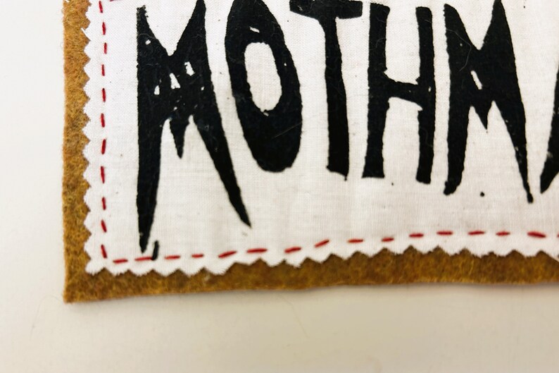 以下が含まれることがあります： 白い布のパッチのクローズアップ。黒いステッチと黒い塗料で書かれた「MOTHMAN」という言葉があります。パッチは、縁に赤いステッチが施された茶色の革の裏地に縫い付けられています。