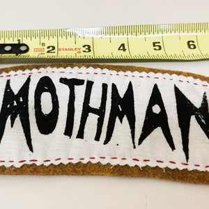 以下が含まれることがあります： 黒い塗料で「MOTHMAN」と書かれた白い布のパッチ。パッチは赤いステッチで茶色のフェルトの裏地に縫い付けられています。