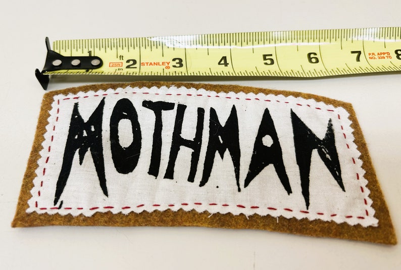 以下が含まれることがあります： 黒い塗料で「MOTHMAN」と書かれた白い布のパッチ。パッチは赤いステッチで茶色のフェルトの裏地に縫い付けられています。