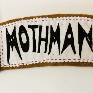以下が含まれることがあります： 白い布の中央に茶色のフェルトパッチがあります。布の中央には黒い文字で「MOTHMAN」と書かれています。