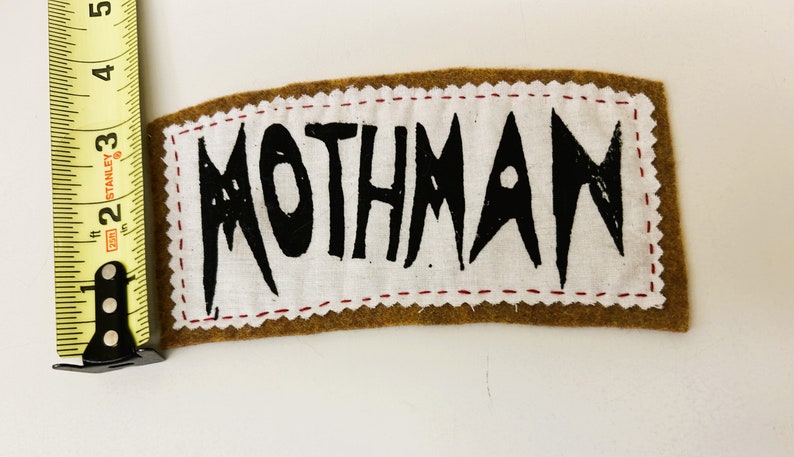 以下が含まれることがあります： 白い布の中央に茶色のフェルトパッチがあります。布の中央には黒い文字で「MOTHMAN」と書かれています。