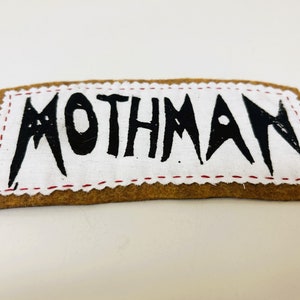 May include: A brown felt patch with red stitching around the edges. The patch has a white background with the word "MOTHMAN" written in black.