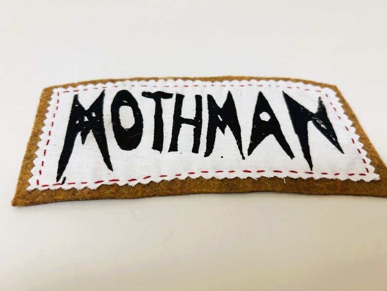 以下が含まれることがあります： 縁が赤いステッチで縫われた茶色のフェルトパッチ。パッチは白地に黒で「MOTHMAN」と書かれています。