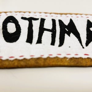 以下が含まれることがあります： 茶色の縁取りと赤いステッチが付いた白い布のパッチ。パッチには黒い塗料で「MOTHMAN」と書かれています。
