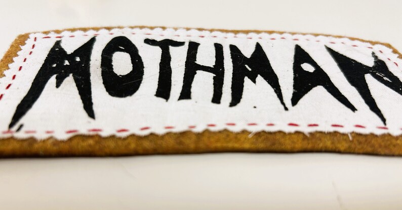 以下が含まれることがあります： 茶色の縁取りと赤いステッチが付いた白い布のパッチ。パッチには黒い塗料で「MOTHMAN」と書かれています。