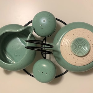 May include: A green ceramic teapot with a white and green lid, a matching creamer, and two salt and pepper shakers, all on a black metal stand.