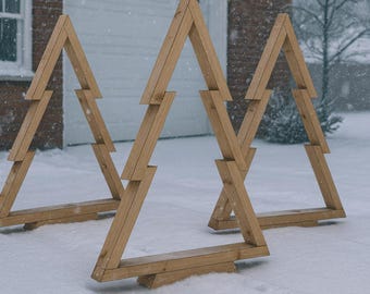 May include: Three wooden Christmas tree decorations stand in the snow. The trees are constructed from light-colored wood and have a geometric design. A brick building and a garage door are in the background.