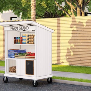 May include: A white mobile farm stand with a slanted roof and the words "FARM STAND" across the top. The stand has shelves stocked with various packaged goods, produce, and firewood. It is positioned on a sidewalk next to a grassy area.
