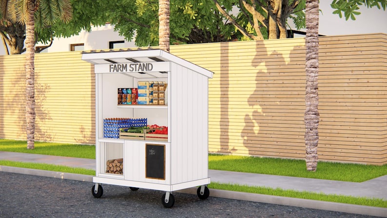 May include: A white mobile farm stand with a slanted roof and the words "FARM STAND" across the top. The stand has shelves stocked with various packaged goods, produce, and firewood. It is positioned on a sidewalk next to a grassy area.