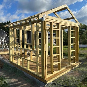 K&ouml;nnte beinhalten: Ein teilweise konstruierter Holzschuppenrahmen, der die St&auml;nder, Sparren und das Fundament der Struktur zeigt. Das Naturholz steht vor dem Hintergrund eines bew&ouml;lkten blauen Himmels und gr&uuml;nen Grases, mit einer Scheune und einer Leiter im Hintergrund.
