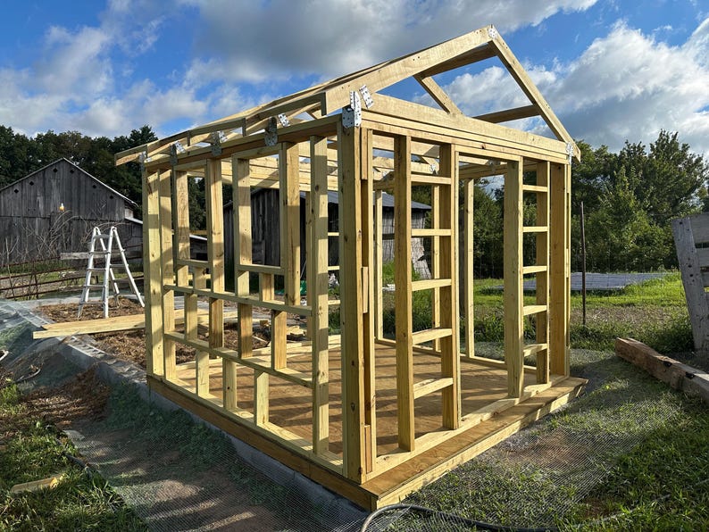 K&ouml;nnte beinhalten: Ein teilweise konstruierter Holzschuppenrahmen, der die St&auml;nder, Sparren und das Fundament der Struktur zeigt. Das Naturholz steht vor dem Hintergrund eines bew&ouml;lkten blauen Himmels und gr&uuml;nen Grases, mit einer Scheune und einer Leiter im Hintergrund.