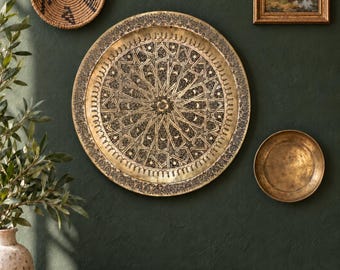May include: A large circular brass tray featuring an intricate engraved geometric pattern with star and crescent motifs. The tray is displayed alongside a small woven basket, a framed landscape painting, and a plain brass bowl on a dark wall.