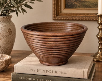 Puede incluir: Un cuenco circular de color marrón oscuro con una superficie exterior estriada y texturizada. El cuenco está colocado sobre dos libros apilados, uno titulado The KINFOLK Home y el otro AT HOME WITH PLANTS, sobre una superficie de madera.