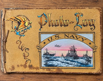 Puede incluir: Un álbum de fotos de ante color canela con el texto Photo-Log y U.S. Navy. La cubierta muestra un gráfico de un águila posada sobre un ancla, un diseño de cuerda y una ilustración rectangular de barcos navales en el mar con un avión.