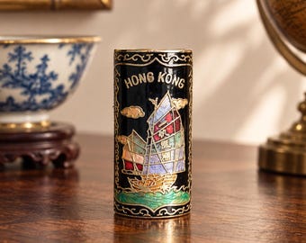 Puede incluir: Un recipiente cilíndrico de metal negro con un diseño de esmalte cloisonné colorido que muestra un barco junco tradicional navegando sobre olas verdes. El texto HONG KONG está impreso en letras doradas en la parte superior, con bordes de filigrana.