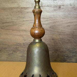 May include: A brass bell with a wooden handle featuring a turned design and a brass finial at the top. The bell has a flared base with a series of small, cut-out shapes along the bottom edge, showing a weathered, metallic patina throughout the surface.