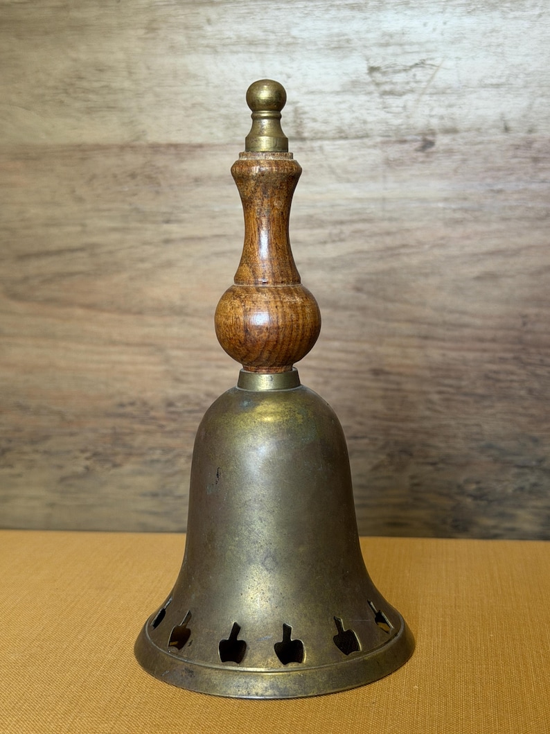 May include: A brass bell with a wooden handle featuring a turned design and a brass finial at the top. The bell has a flared base with a series of small, cut-out shapes along the bottom edge, showing a weathered, metallic patina throughout the surface.