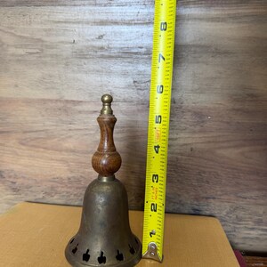 May include: A brass bell with a wooden handle and a row of small apple-shaped cutouts along the bottom edge. A yellow measuring tape placed next to the bell indicates a total height of approximately 9 inches from the base to the top finial.