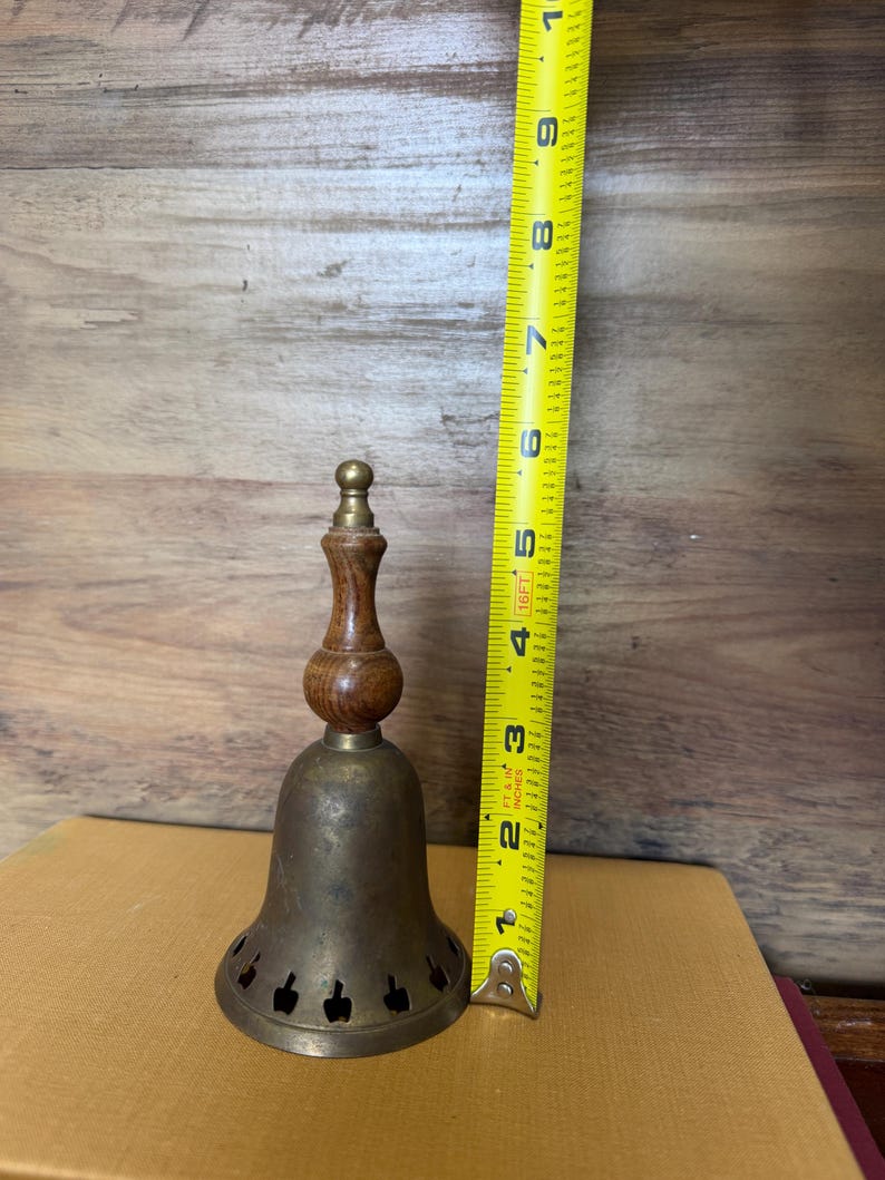 May include: A brass bell with a wooden handle and a row of small apple-shaped cutouts along the bottom edge. A yellow measuring tape placed next to the bell indicates a total height of approximately 9 inches from the base to the top finial.
