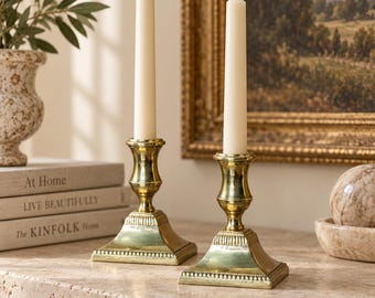 Puede incluir: Dos candelabros de latón pulido con bases cuadradas y detalles de cuentas, cada uno con una vela cónica de color blanco roto, alta y delgada. Los candelabros de metal presentan un acabado liso y reflectante y un diseño clásico.
