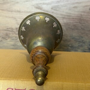 May include: A brass bell with a wooden handle and a flared base featuring a series of small, cut-out shapes along the rim. The bell has a metallic, aged finish and is positioned vertically, showing the handle and the decorative perforated edge.