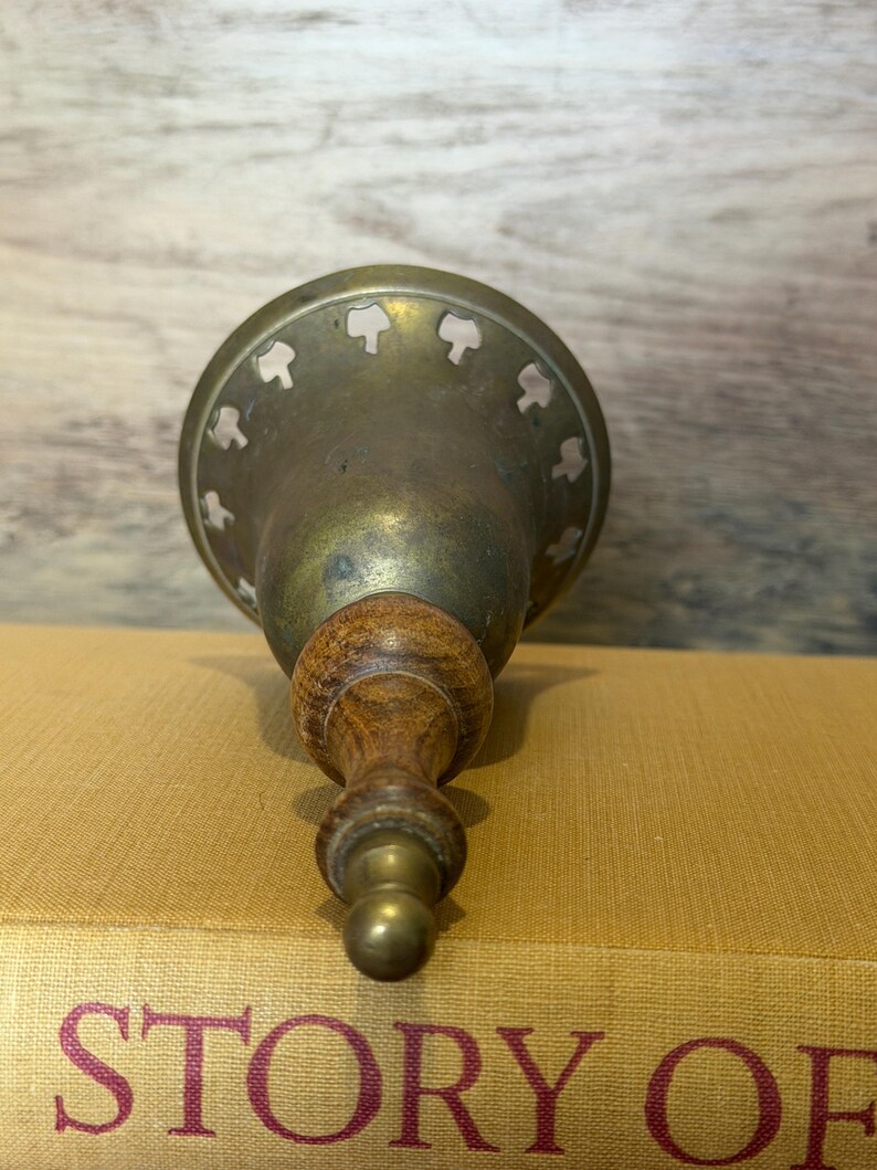 May include: A brass bell with a wooden handle and a flared base featuring a series of small, cut-out shapes along the rim. The bell has a metallic, aged finish and is positioned vertically, showing the handle and the decorative perforated edge.