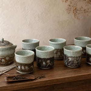 May include: A ceramic tea set consisting of a lidded teapot and six matching cups. Each piece features a pale green crackle glaze on the upper portion and a dark brown, textured base with cutout details. A small wooden scoop holds loose tea leaves.